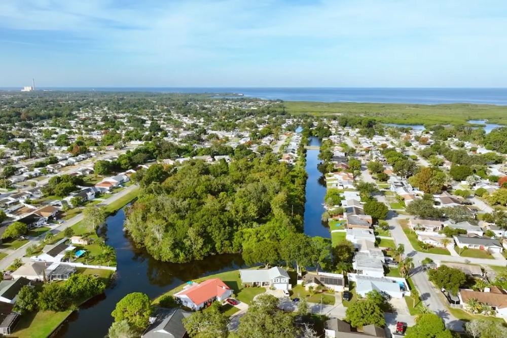 Aerial view of a coastal suburb with tree-lined canals flanked by houses, fading into the sea on the horizon.