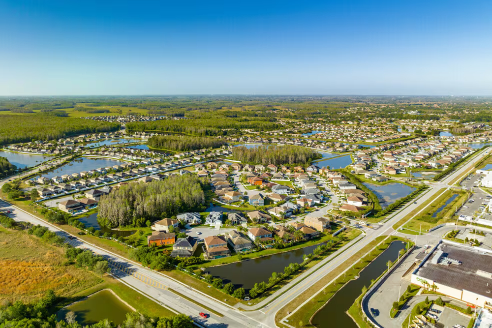 Aerial view of a suburban neighborhood with winding canals, ponds, and grid-like streets surrounded by green spaces under a clear blue sky, far from the city center.