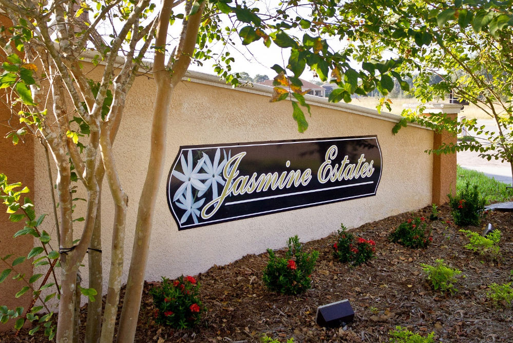 Jasmine Estates Exterior sign reading 'Jasmine Estates' on a beige stucco wall, with trees and a landscaped garden in the foreground